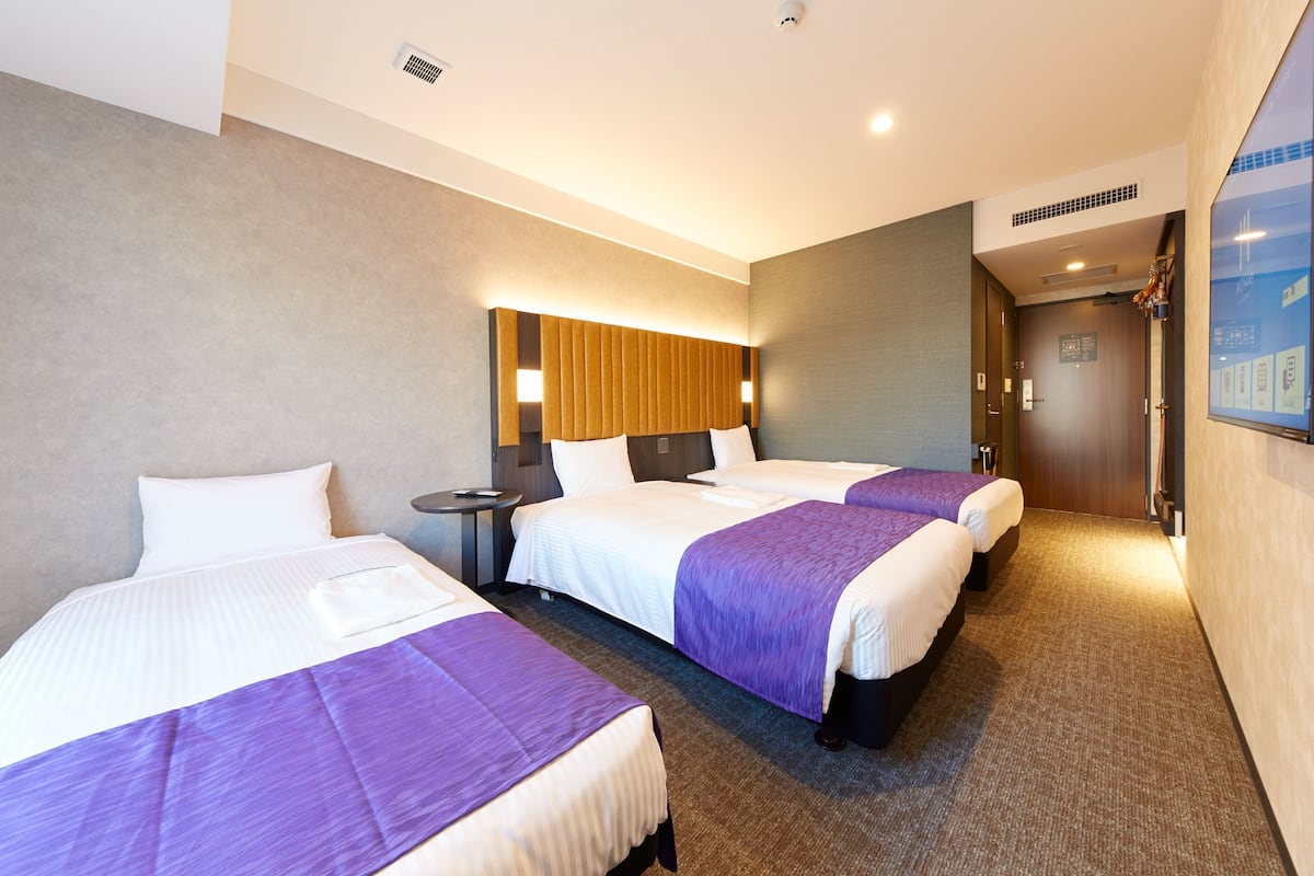 A hotel room with three beds, a small table, and a wall-mounted TV.