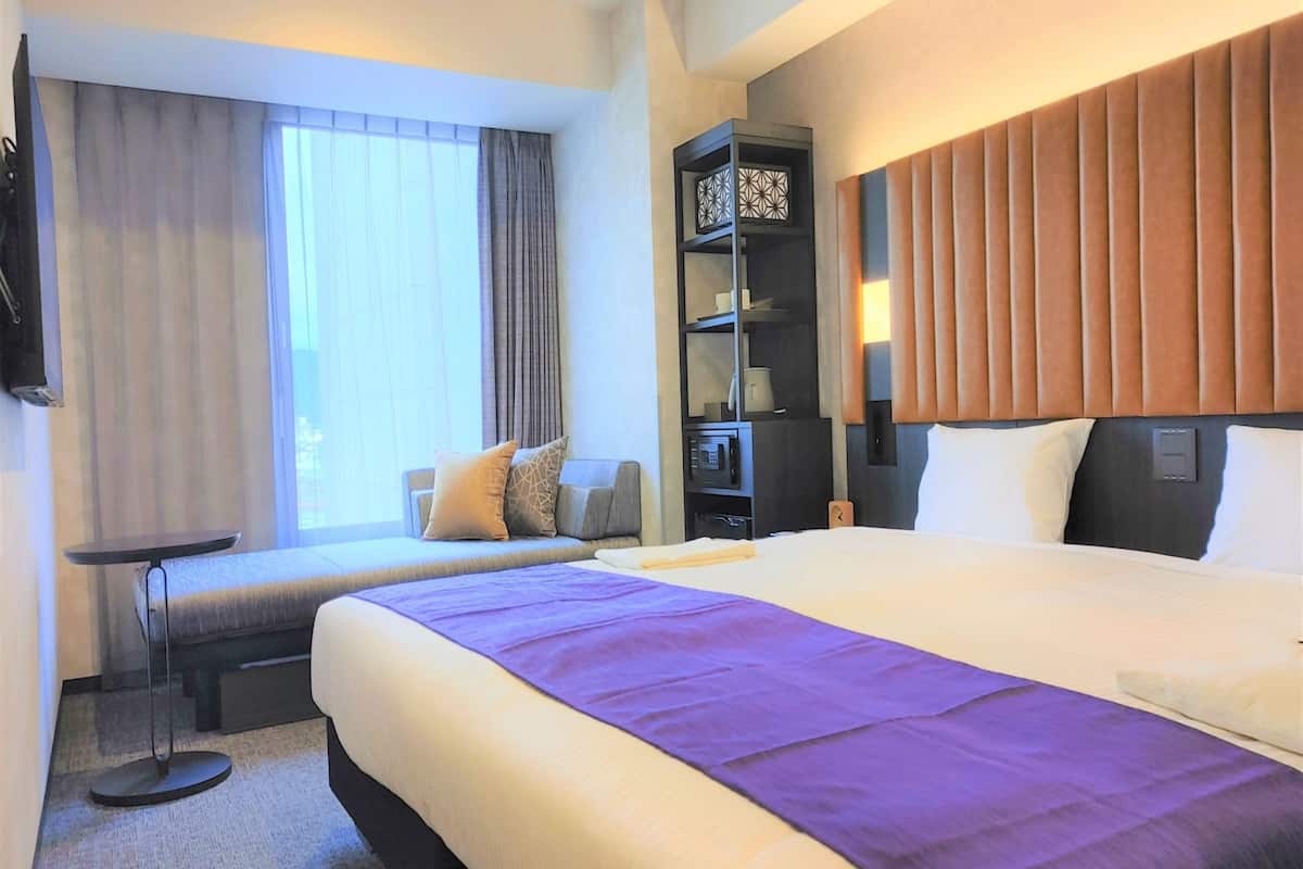 A hotel room with a large bed, a seating area with a bench, a TV mounted on the wall, and a bookshelf.