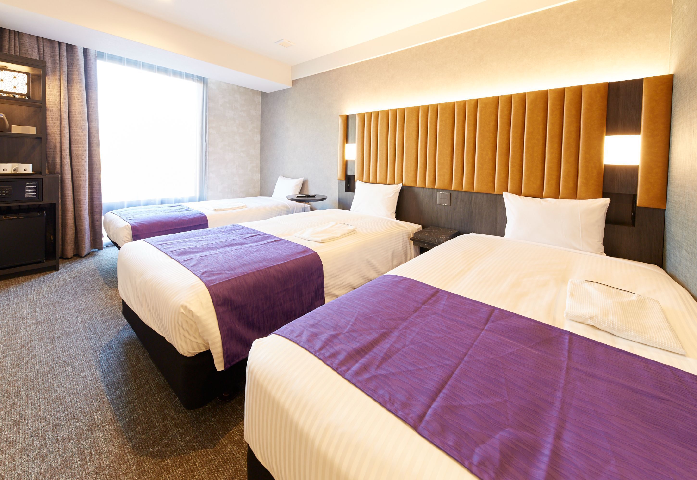 Habitación de hotel con tres camas, cada una con una colcha morada y ropa blanca. Hay una mesita de noche y una lámpara entre las camas, y un ventanal con cortinas.