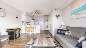 Una sala de estar moderna con un sofá gris, una mesa de centro de madera y un tapete colorido. En el fondo, se ve una cocina con armarios blancos y un comedor con una mesa y sillas de madera.