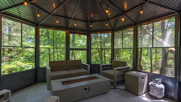 A conservatory with a built-in fire pit, seating area, and large windows offering forest views.