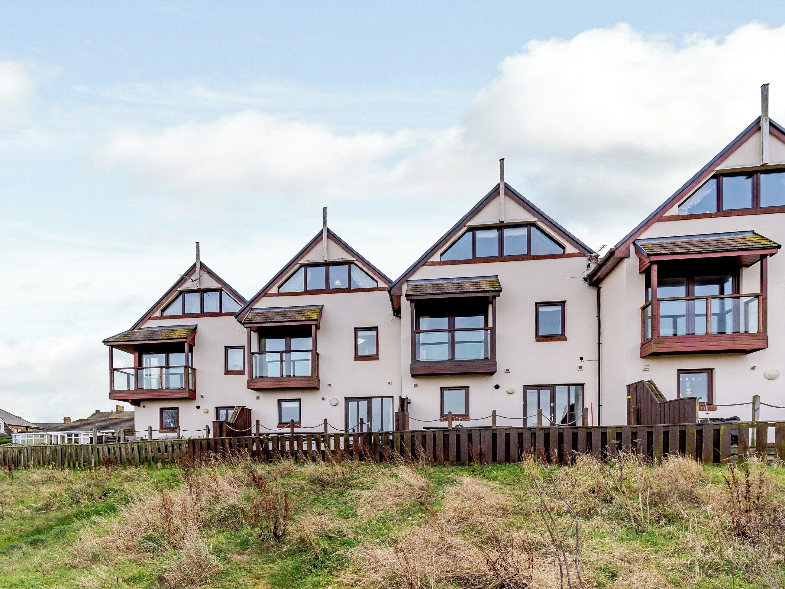 A row of three-story houses with multiple windows and balconies, surrounded by a wooden fence and grassy area.