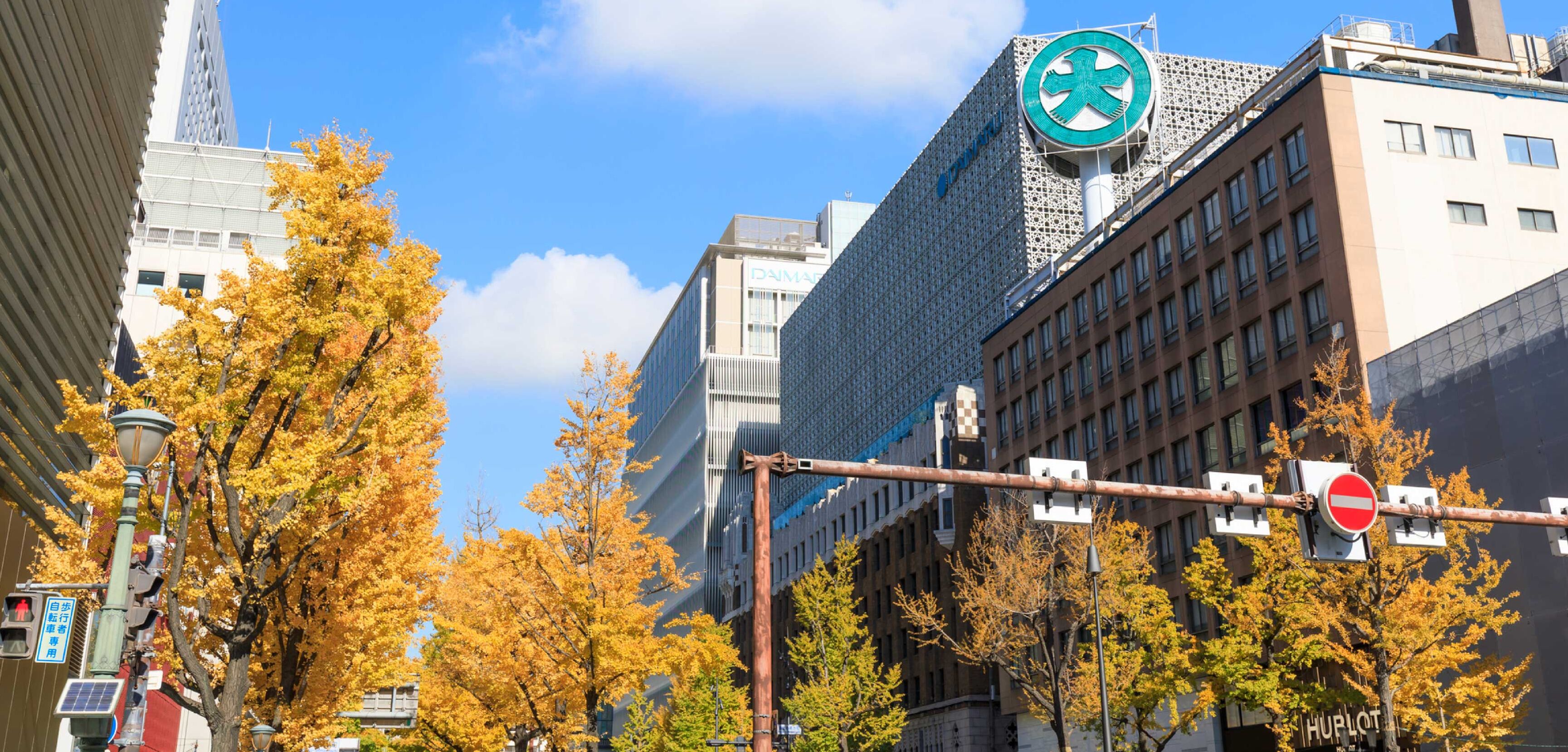 Ein Gebäude mit einem großen, runden Logo an der Fassade, umgeben von Herbstbäumen und moderner Architektur.