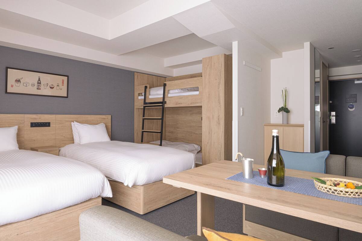 A hotel room with two beds, a wooden desk, a small table, a bottle of wine, and a framed picture on the wall.