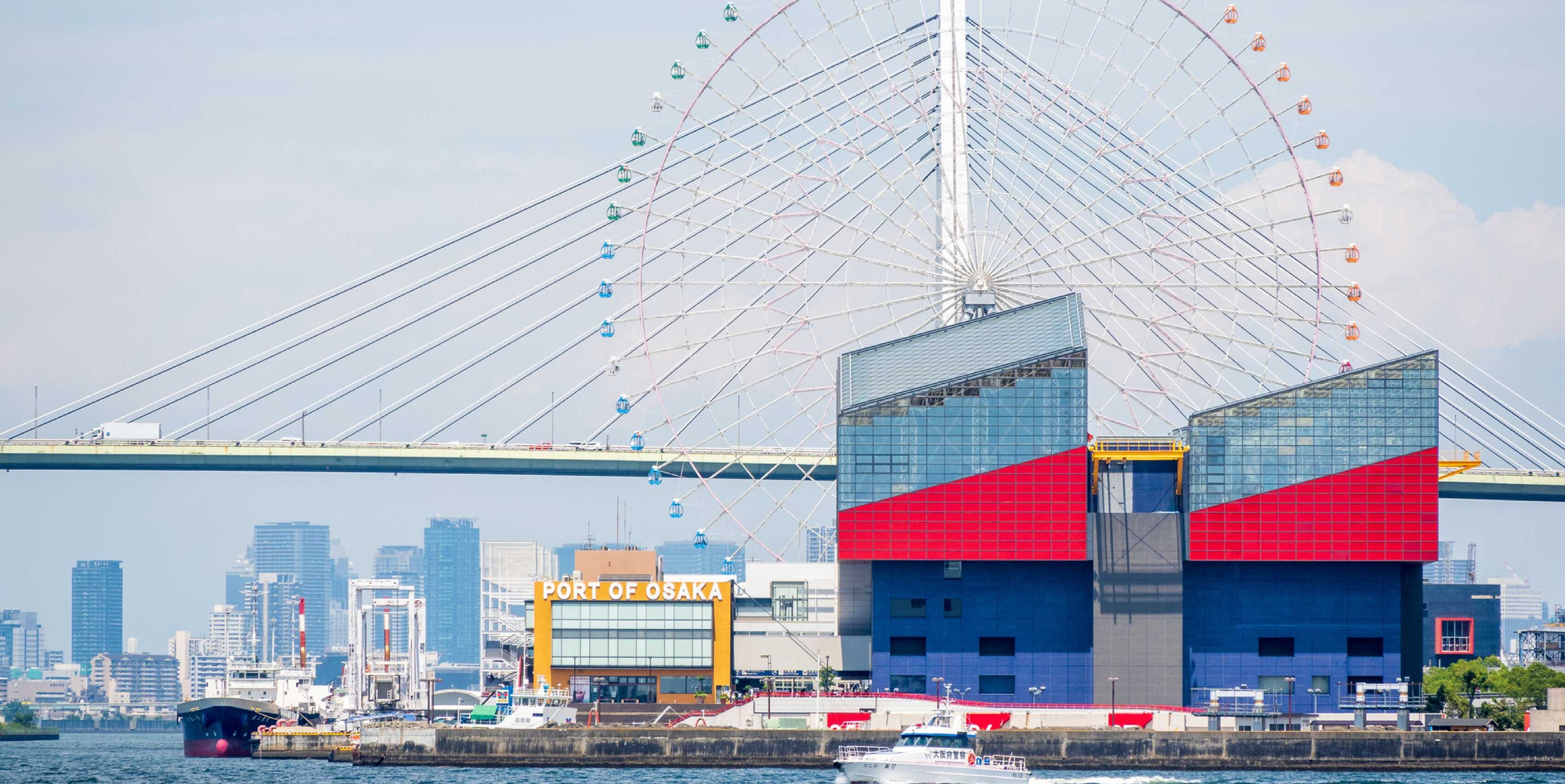 A modern building with a prominent red and blue facade, located near a waterfront with a large bridge in the background.
