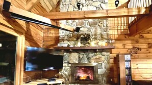 Ein gemütliches Blockhäuschen-Interieur mit Kamin aus Stein, Holzwänden, einem Flachbildfernseher und einer Bücherwand.