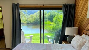 Un dormitorio con una cama grande, una mesita de noche con lámpara y vistas a un lago y vegetación.