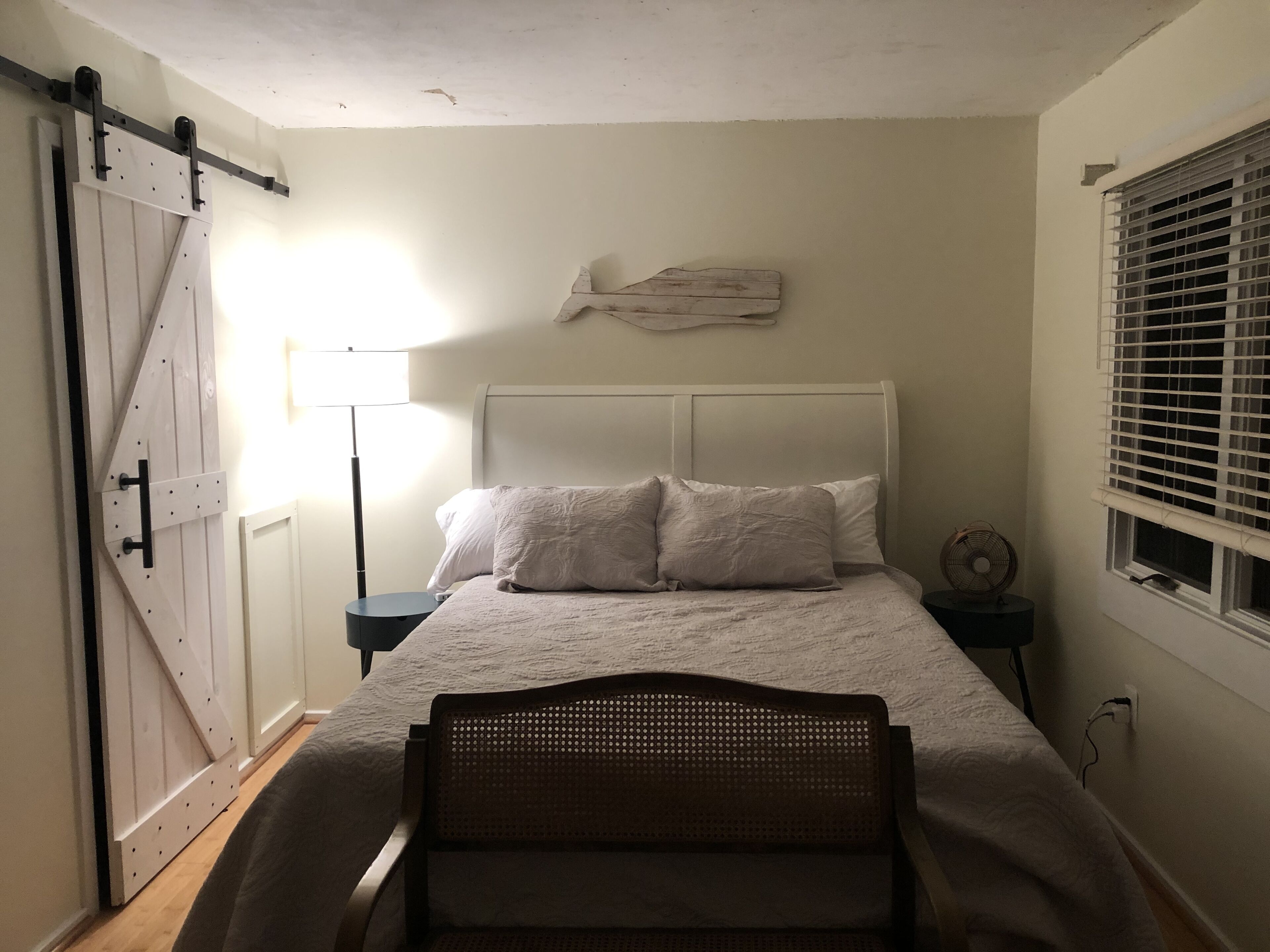 Ein Schlafzimmer mit einem Bett, einem Nachttisch, einer Lampe, einem Ventilator, einem Fenster mit Jalousien und einer Schiebetür.