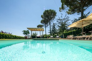 A clear swimming pool with lounge chairs and umbrellas, surrounded by trees and a pergola.