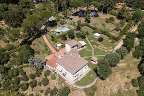 An aerial view of a property with a large house, a swimming pool, and surrounding greenery.