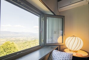 A window with a view of a landscape, a lamp, and a pillow on a bedside table.