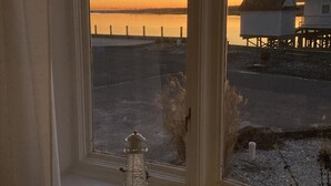 A view through a window with a white lamp on the sill, overlooking a calm body of water and a shoreline with buildings and a fence. - Bayfront Bungalow (Cape May Court House)