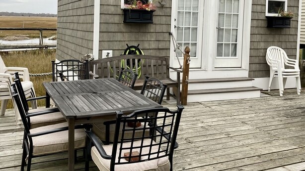 A wooden deck with a table and chairs, a small shed with windows and a door, and a view of the landscape. - Bayfront Bungalow (Cape May Court House)