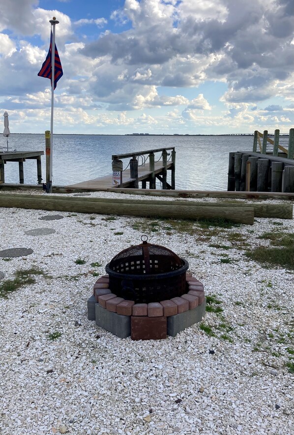 A pebbled beach area with a fire pit, a dock, and a lighthouse in the background.