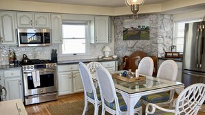 A kitchen with white cabinets, stainless steel appliances, a dining table with white chairs, and a stone accent wall. - Bayfront Bungalow (Cape May Court House)