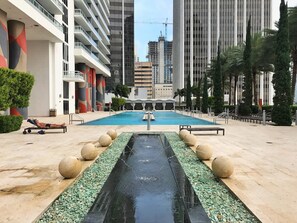 Ein Pool mit Wasserspiel, umgeben von Palmen und modernen Gebäuden.