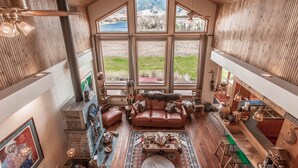 A spacious loft with a large window offering a view of a lake and mountains, a leather sofa, a fireplace, and a wooden ceiling. - Cutbow Bend (Livingston)