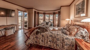 A bedroom with a large bed, wooden headboard, a TV mounted on the wall, and a window with a view of mountains. - Cutbow Bend (Livingston)