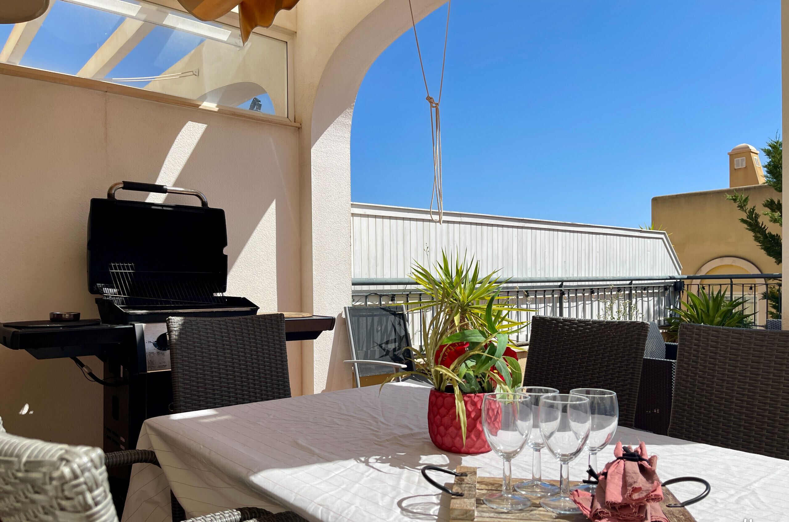 A dining area with a table set for four, a grill, wicker chairs, and a potted plant.