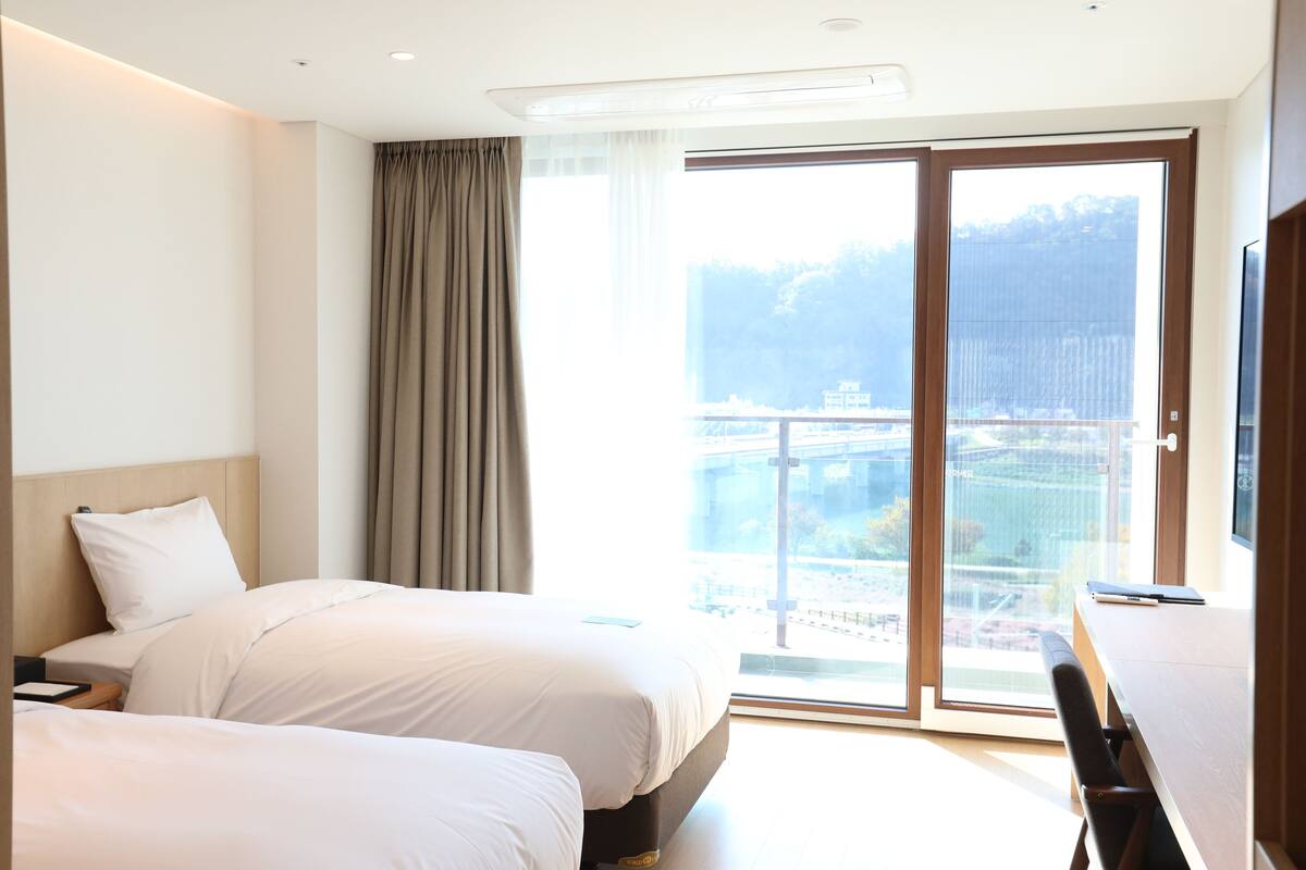 A hotel room with two beds, a desk, and a large window with a view of a cityscape.