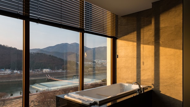 A modern bathroom with a large bathtub, a window with blinds, and a view of a river and mountains. - Hotel Arina (Miryang)