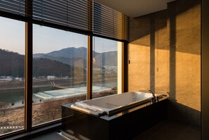 Ein modernes Badezimmer mit einer großen Badewanne, einem Fenster mit Jalousien und Blick auf einen Fluss und Berge.