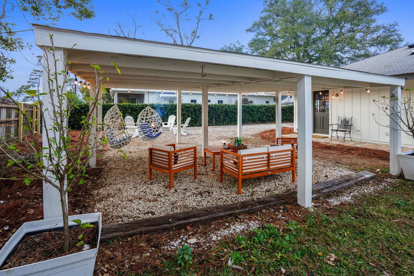 Une terrasse couverte, aménagée avec du mobilier d’extérieur, une pergola et une vue sur une maison en arrière-plan.