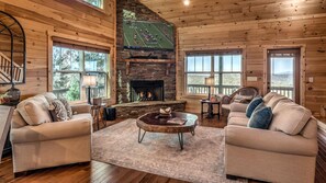 A cozy log cabin interior with a stone fireplace, two sofas, a coffee table, and large windows offering views of the outdoors.