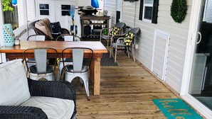 Een overdekt terras met een houten tafel en stoelen, rieten meubels en een gekleurd vloerkleed.