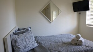 Una cama bien tendida con almohadones estampados, una toalla enrollada, un televisor montado en la pared y una ventana con persianas.