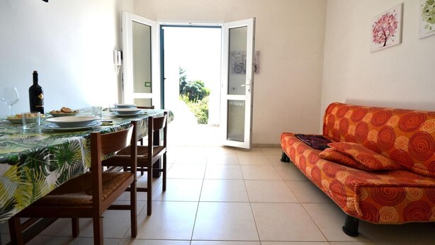 Un comedor con una mesa preparada para cuatro personas, un sofá y una puerta abierta que da al exterior.