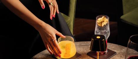 A person seated at a table with a menu, a glass of yellow drink, a glass of red wine, and a small container with a lid.