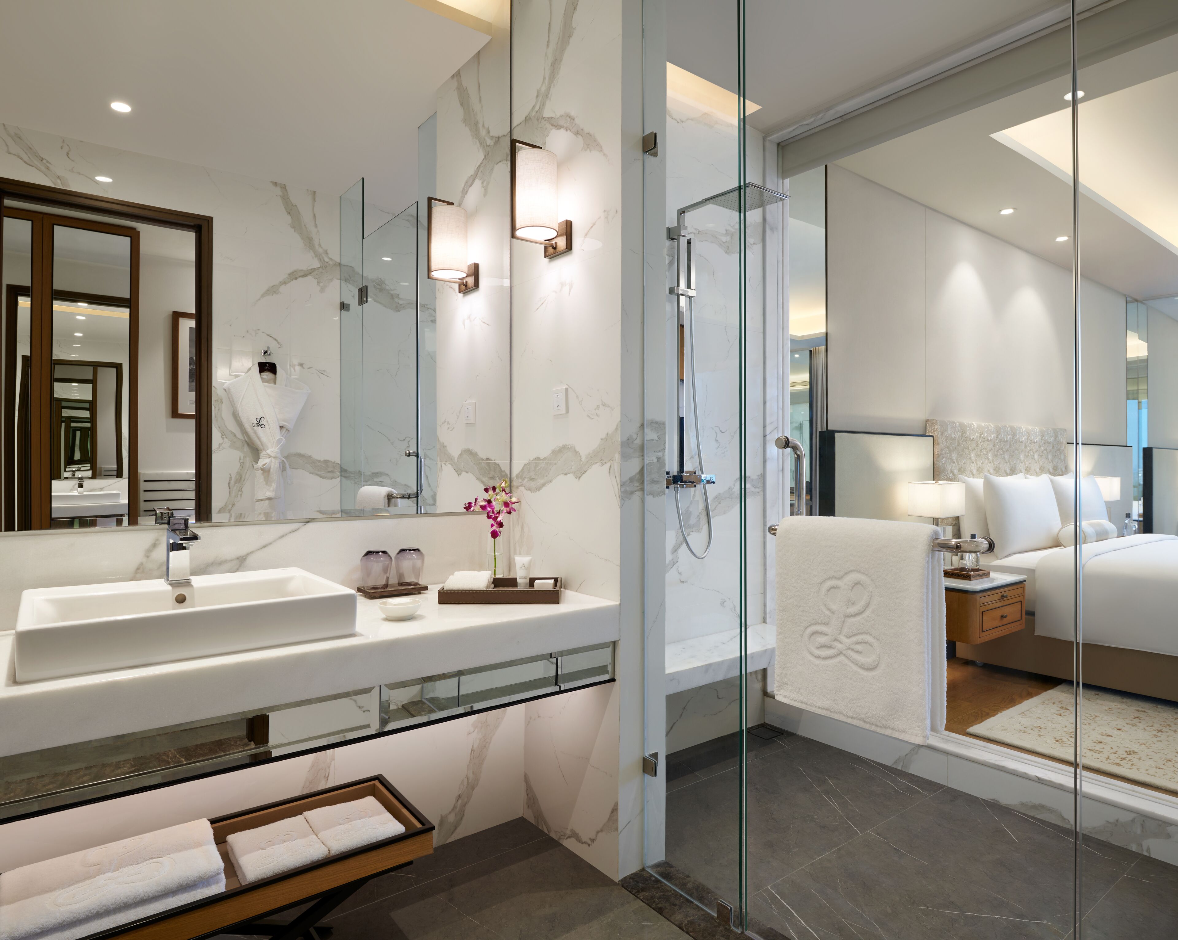 Un baño de hotel moderno con un espejo grande, ducha con cerramiento de vidrio, tocador con encimera de mármol y zona de dormitorio separada con cama y mesita de noche.
