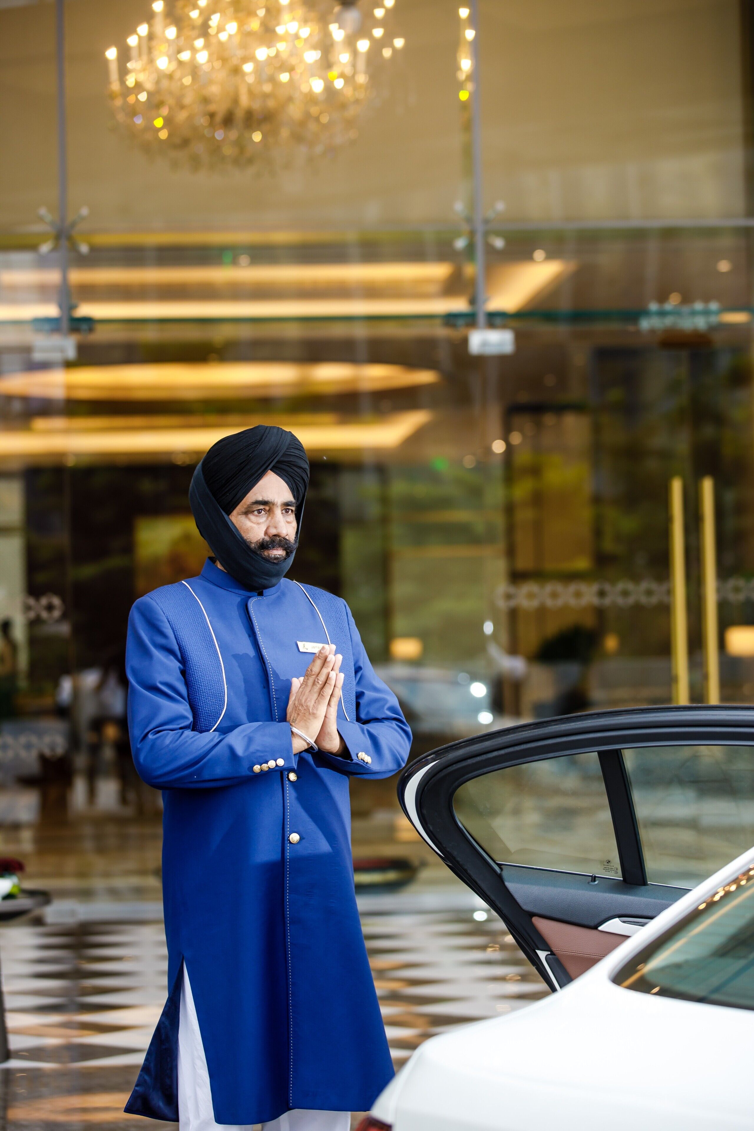 A person in traditional attire standing by a car in front of a building with a glass facade and a chandelier.