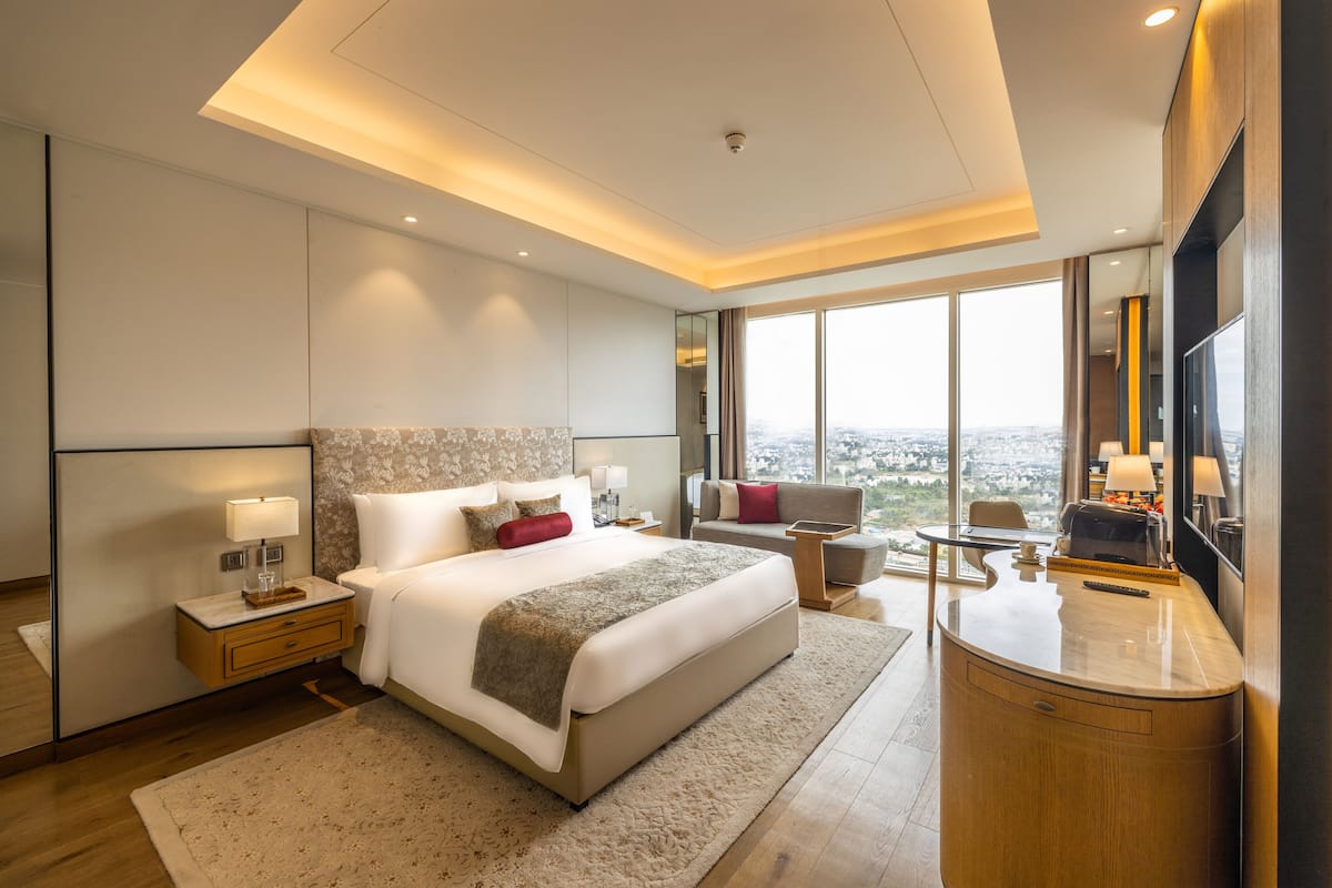 Una habitación de hotel moderna con una cama grande, una zona de estar, un escritorio y un ventanal con vistas a la ciudad.