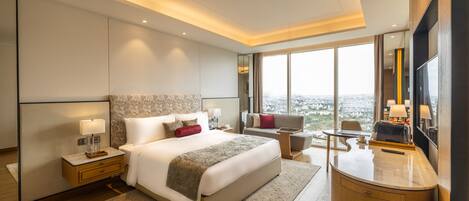 A modern hotel room with a large bed, a sitting area, a desk, and a large window offering a city view.