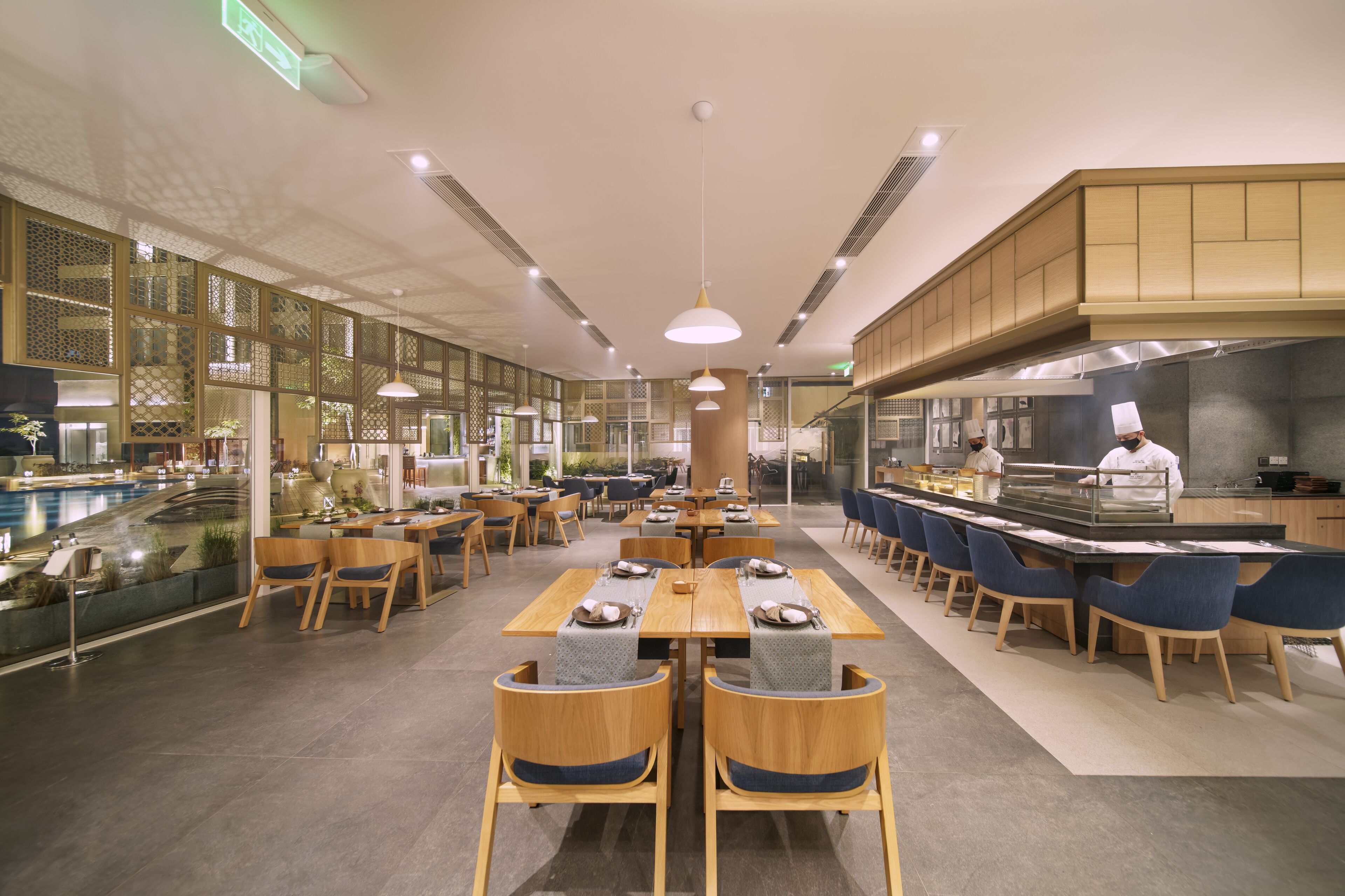A modern restaurant with wooden tables and chairs, a central serving area, and decorative glass partitions.