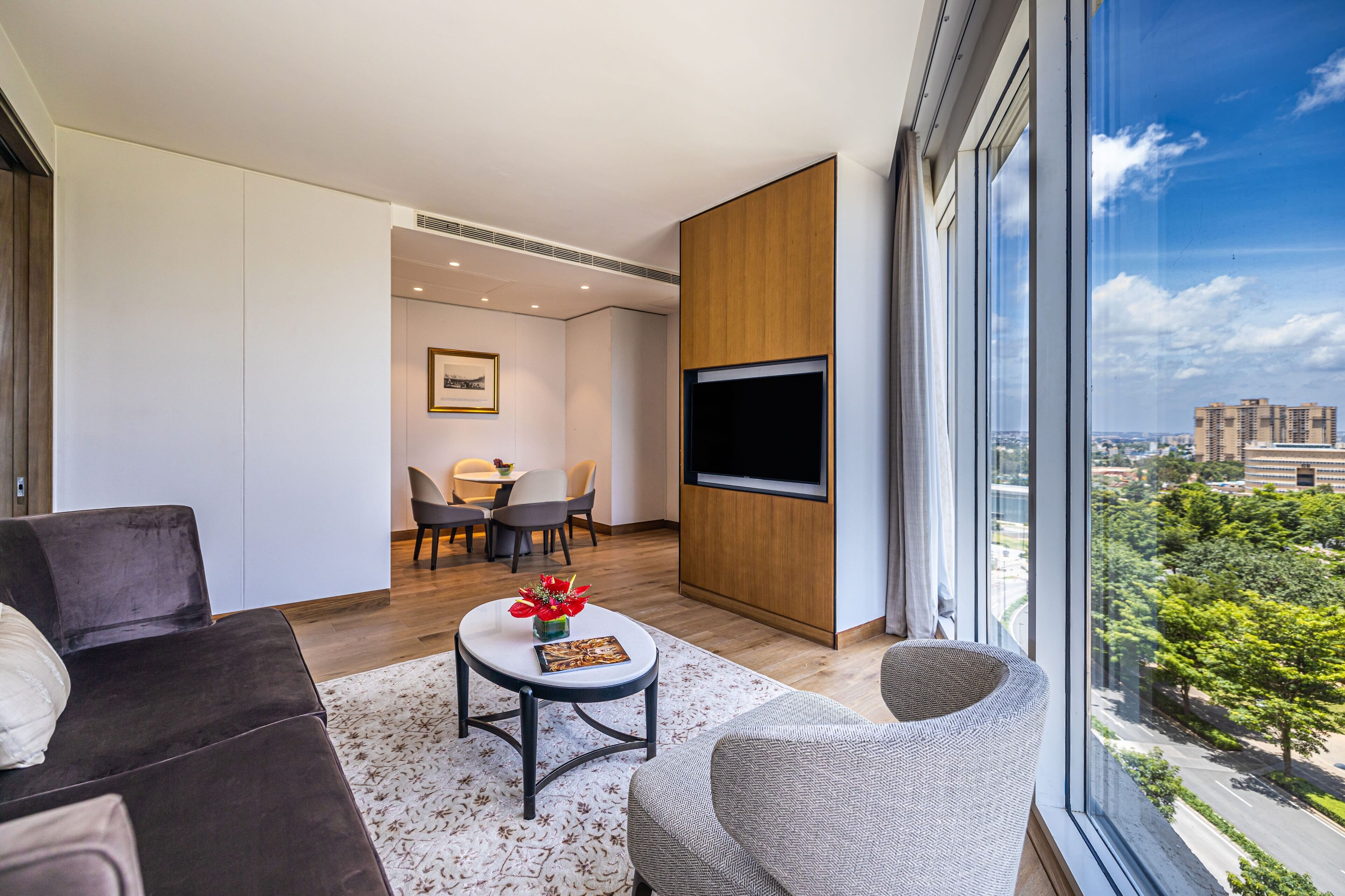 A modern living room with a sofa, armchair, and a coffee table with a vase of flowers. There is a flat-screen TV mounted on the wall and a dining area with chairs and a table.