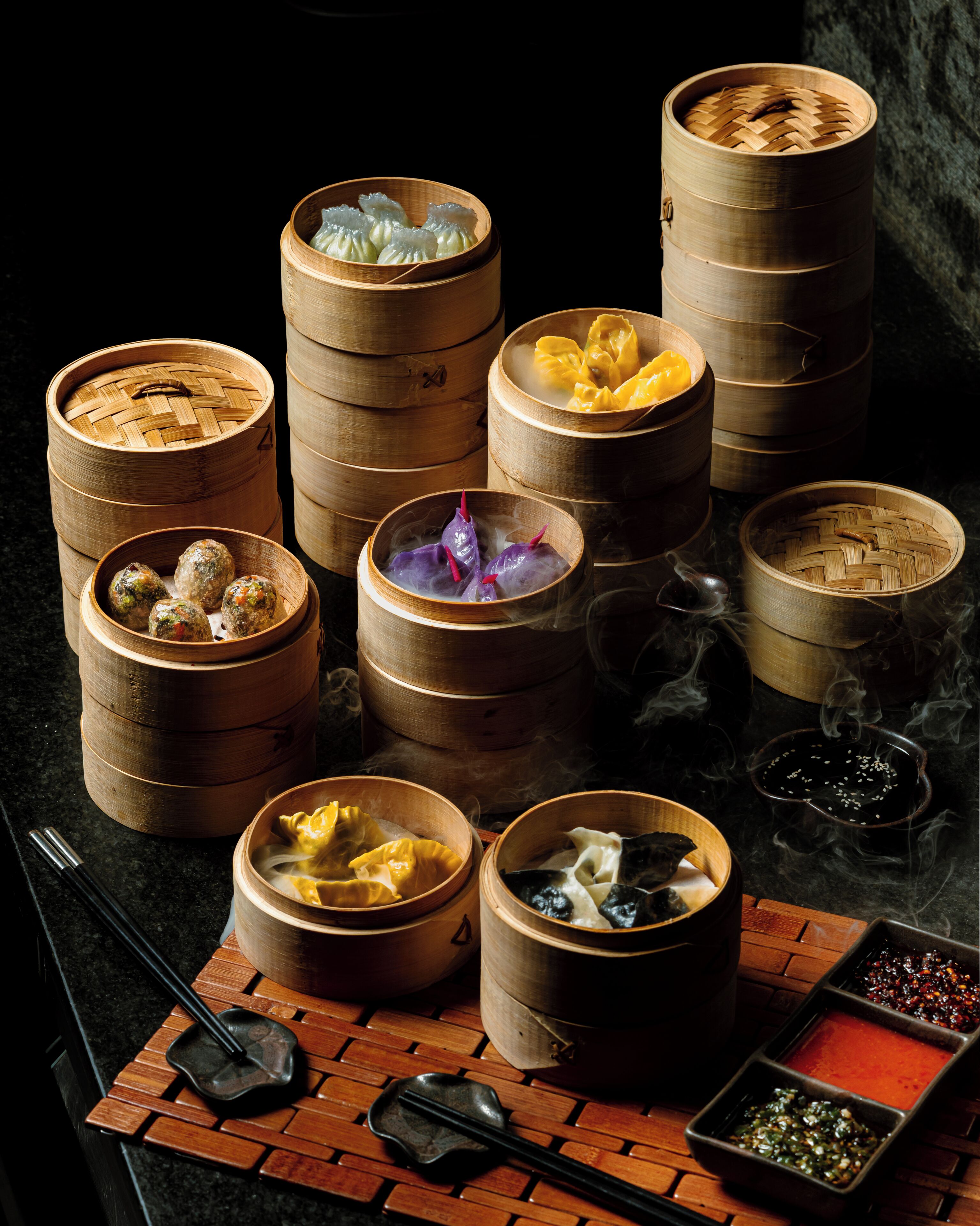 A variety of dim sum dishes in bamboo steamers, a pair of chopsticks, and a small dish of sauces on a wooden table.