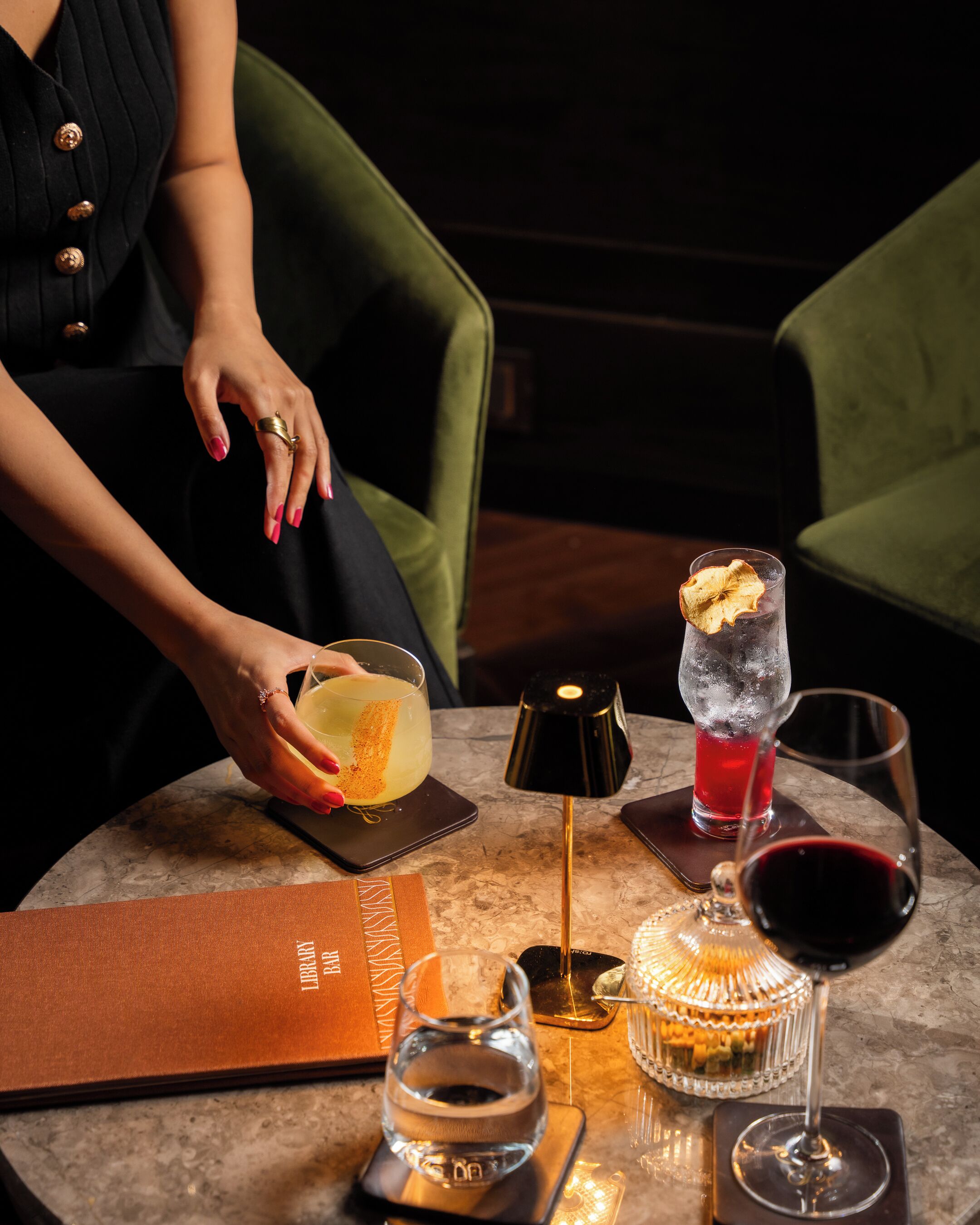 A table with a menu, a glass of red wine, a cocktail with a garnish, a candle, and a water bottle.
