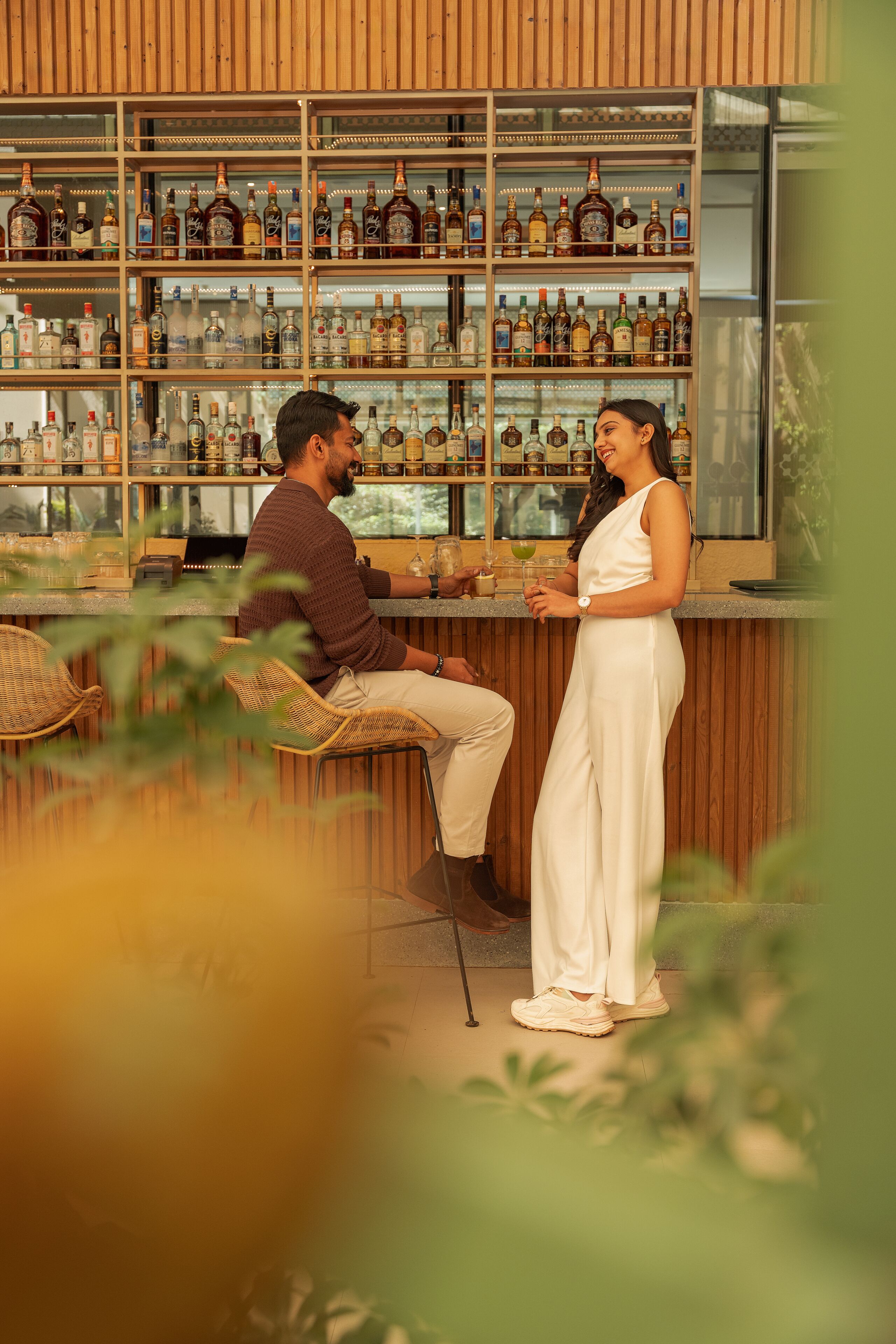 A bar with shelves of bottles, wicker chairs, and a counter where two people are standing.