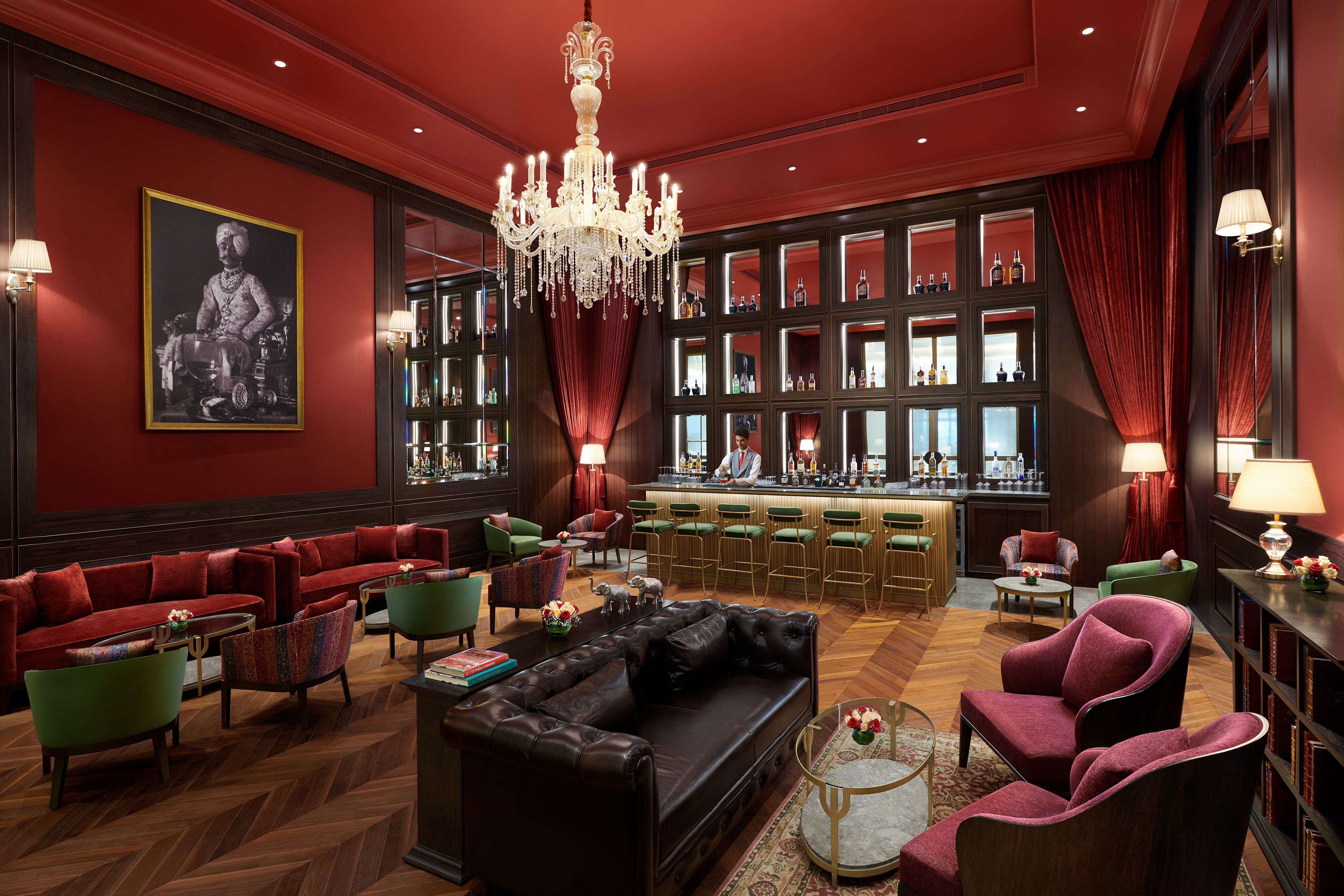 A luxurious lobby with a red color scheme, a large chandelier, a bar area, and a seating area with a black leather sofa and green armchairs.