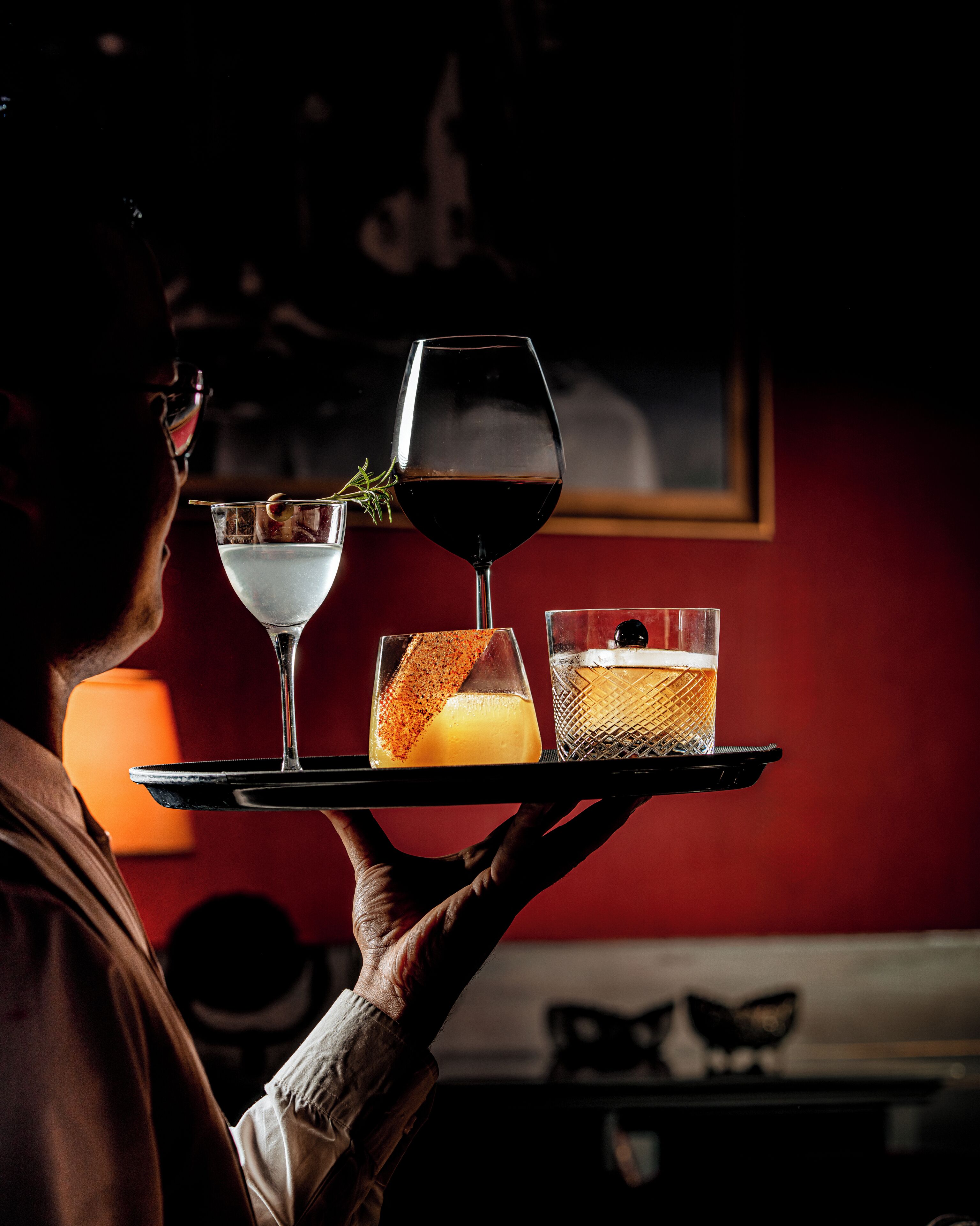 A person holding a tray with a wine glass, a cocktail glass, and a small glass with a garnish.