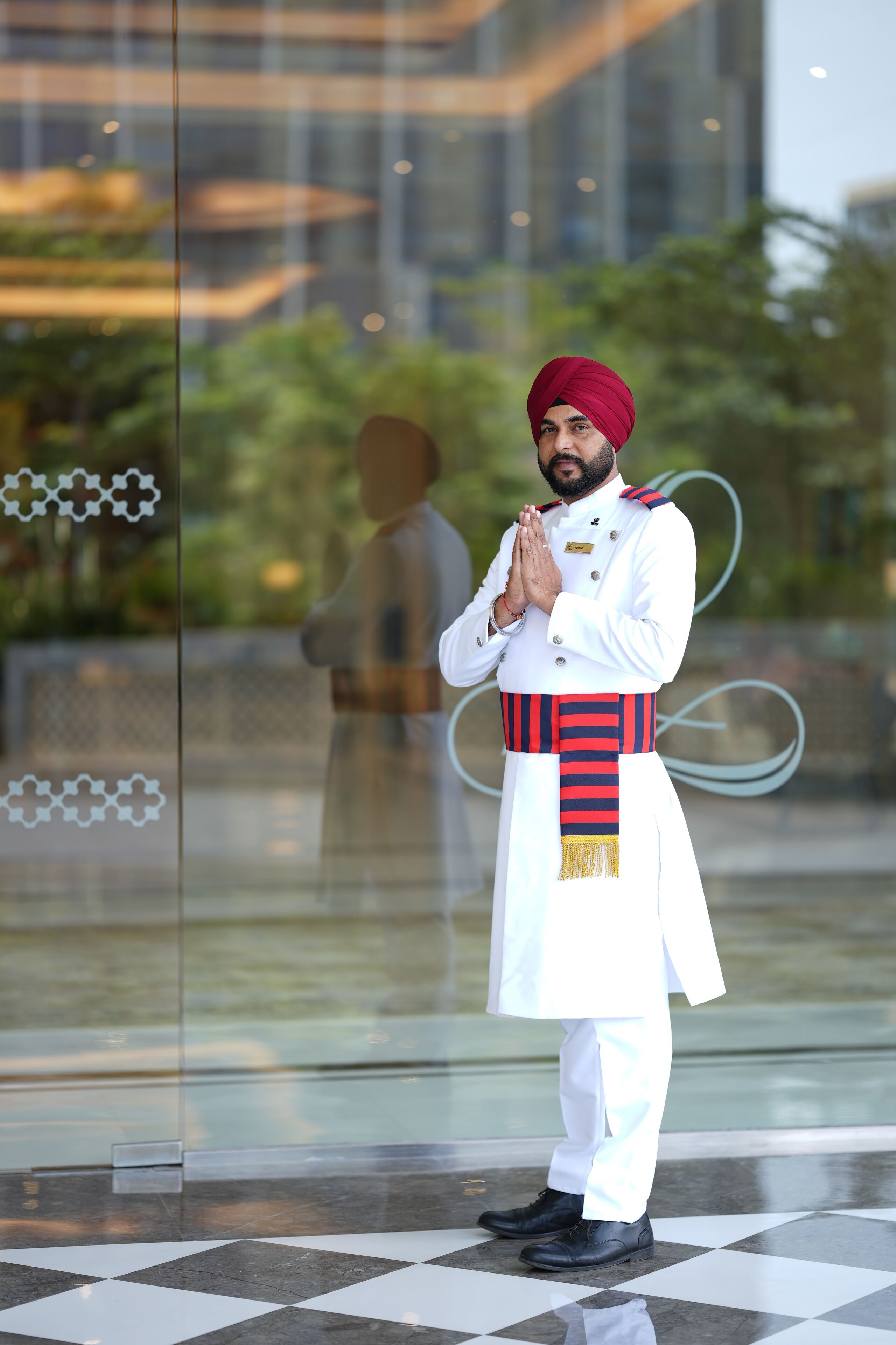 A person in traditional attire standing in front of a glass window with a reflection of greenery and a building.