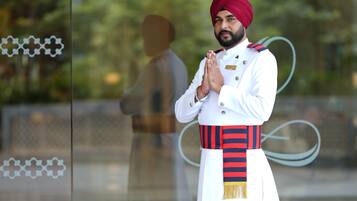 A person in traditional attire standing in front of a glass window with a reflection of greenery and a building.