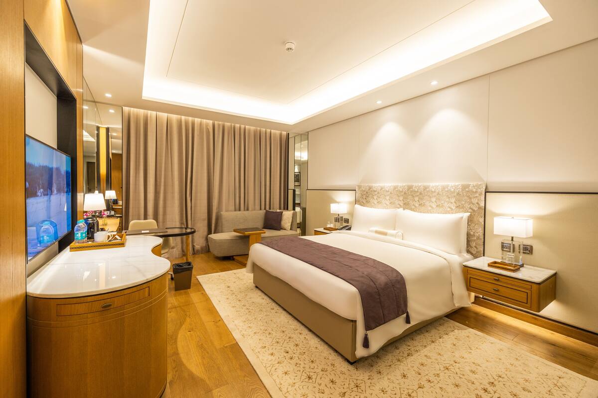 Una habitación de hotel moderna con una cama grande, una zona de estar con sofá y sillones, una pequeña mesa redonda, un escritorio con televisor de pantalla plana y un baño con espejo y toallero.