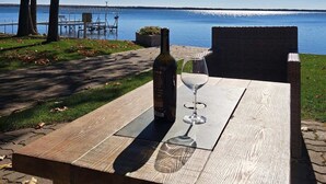 Una mesa de madera con una botella y una copa de vino, sobre un día soleado junto a un lago con árboles y un muelle al fondo.