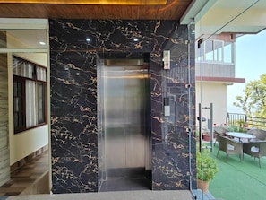 A modern elevator with a marble wall, a glass door, and a small outdoor seating area with a table and chairs. - Hotel Harmony Blue Mcleodganj, Dharamshala (Dharamshala)