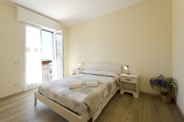 A bedroom with a bed, bedside tables, a nightstand, a lamp, a window, and a view of the outside.