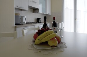 Une cuisine avec des meubles blancs, un four à micro-ondes, une machine à café et une table à manger sur laquelle est disposée une assiette de fruits.